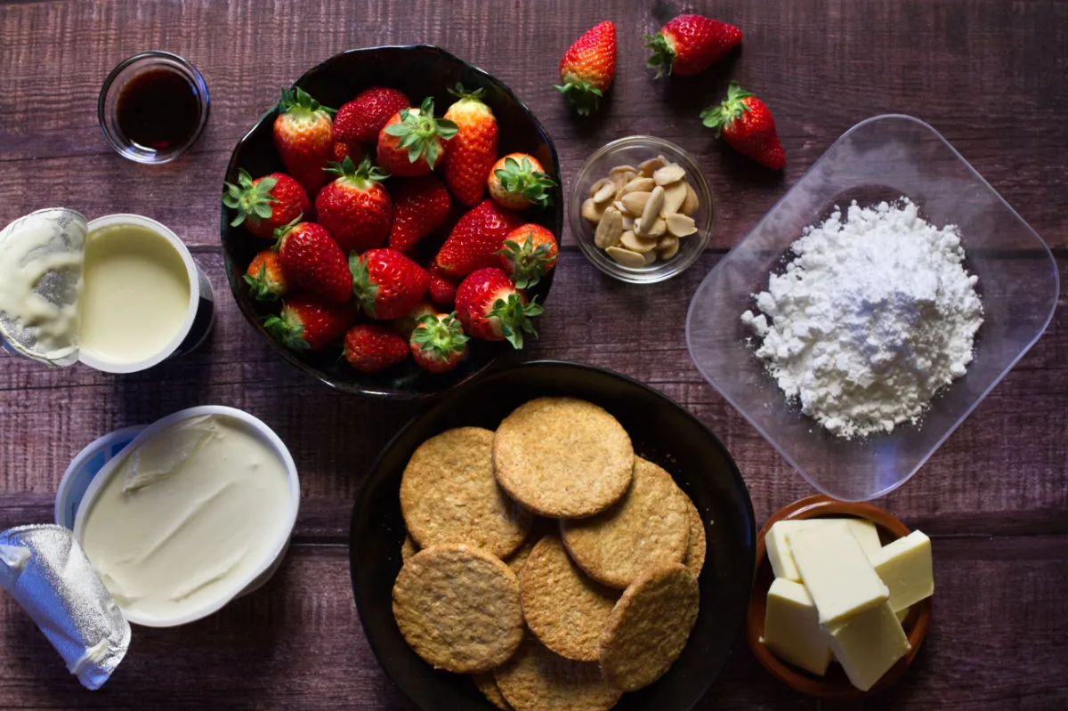 a large bowl of fresh strawberries sists beside some biscuits and other ingredients used to make cheesecake. 