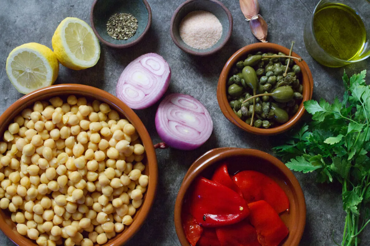 A large bowl of chickpeas sits beside piquillo peppers, capers, red onion, and other veg. 